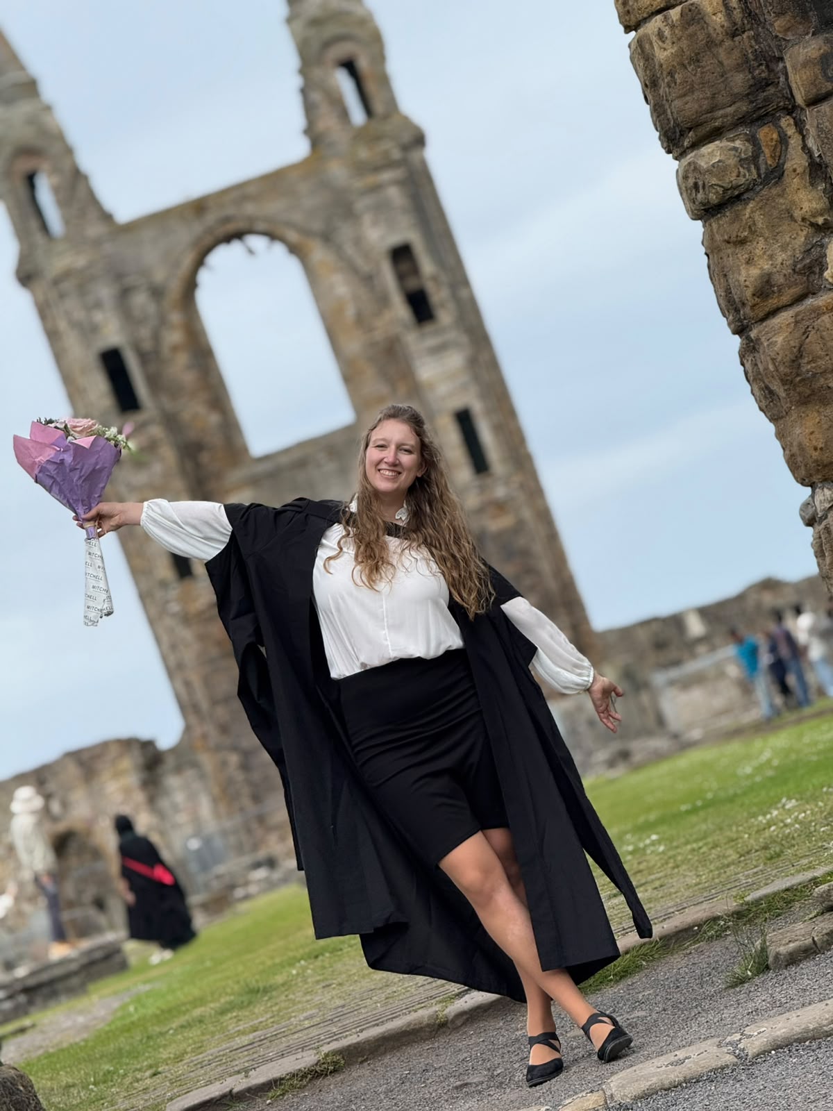 Marta in graduation gown holding flowers at the St Andrews cathedral ruins